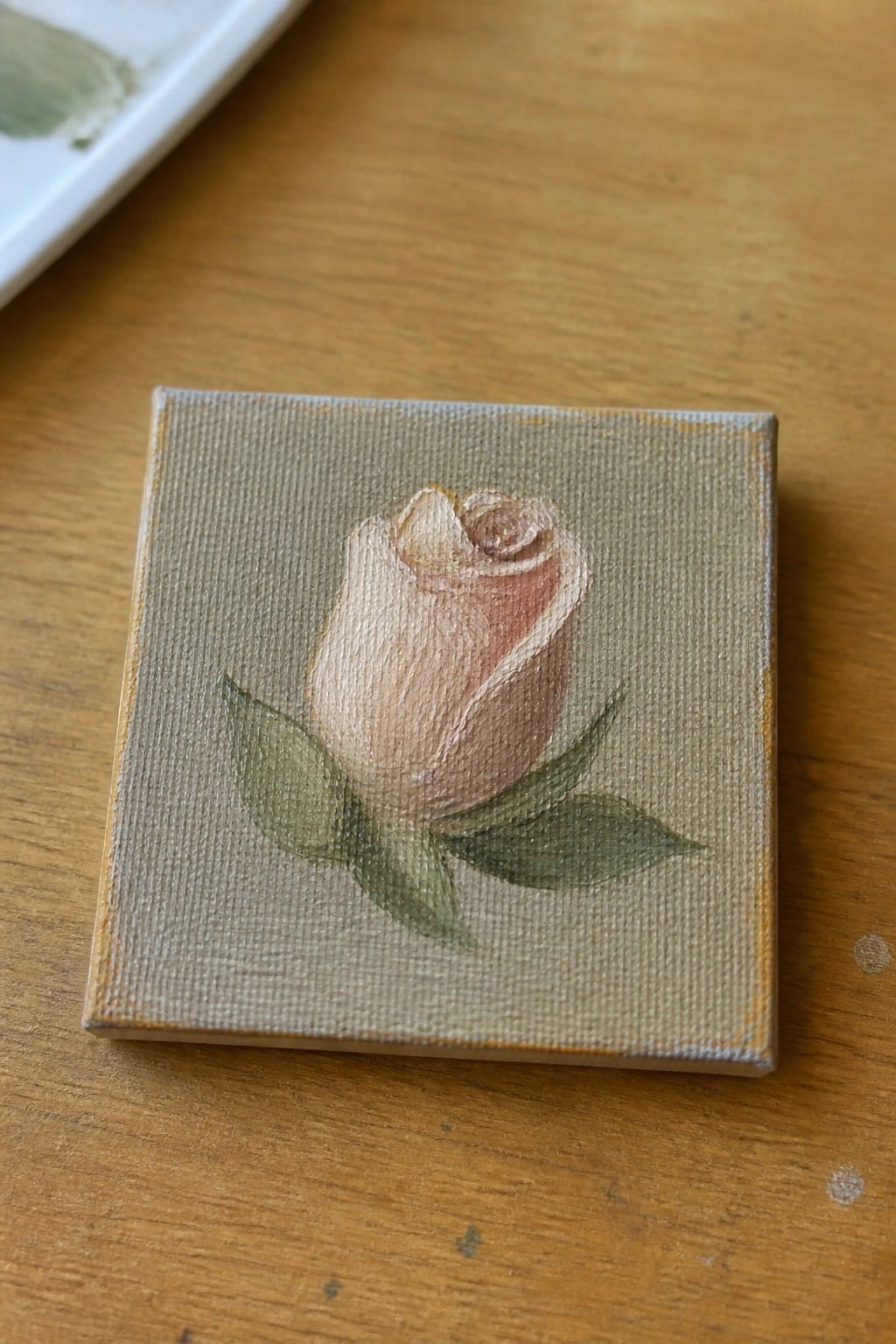 Small canvas with soft pink rosebud and green leaves on wooden table