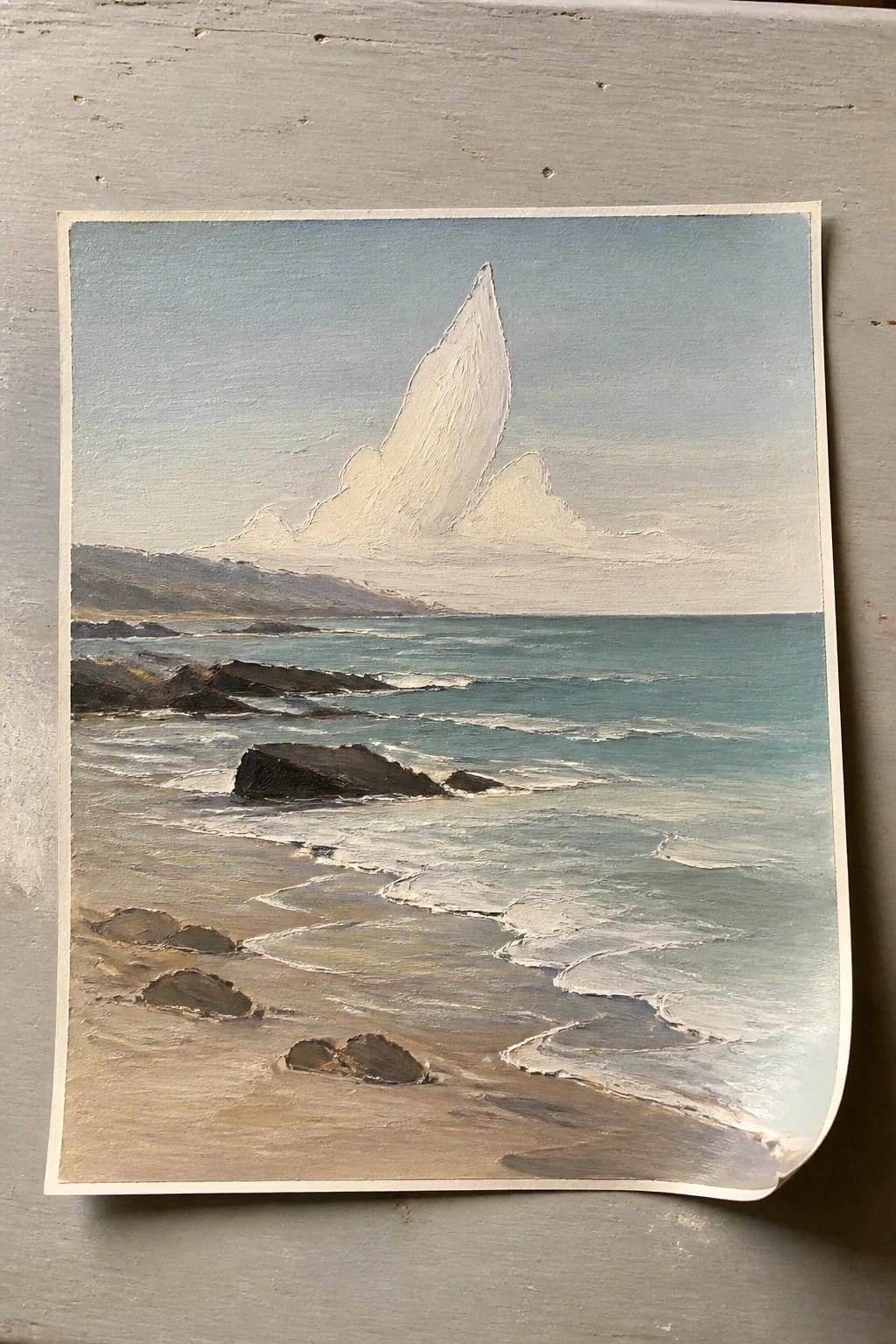 Seascape painting of rocky beach, turquoise waves, distant hills, and large white cloud.