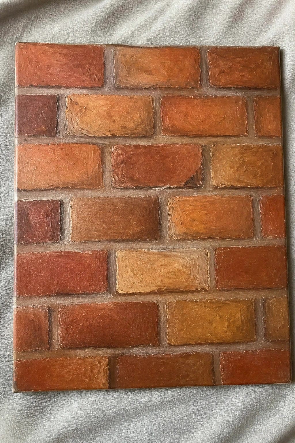 Rectangular painting of red, orange, and brown bricks forming a textured wall on gray fabric.