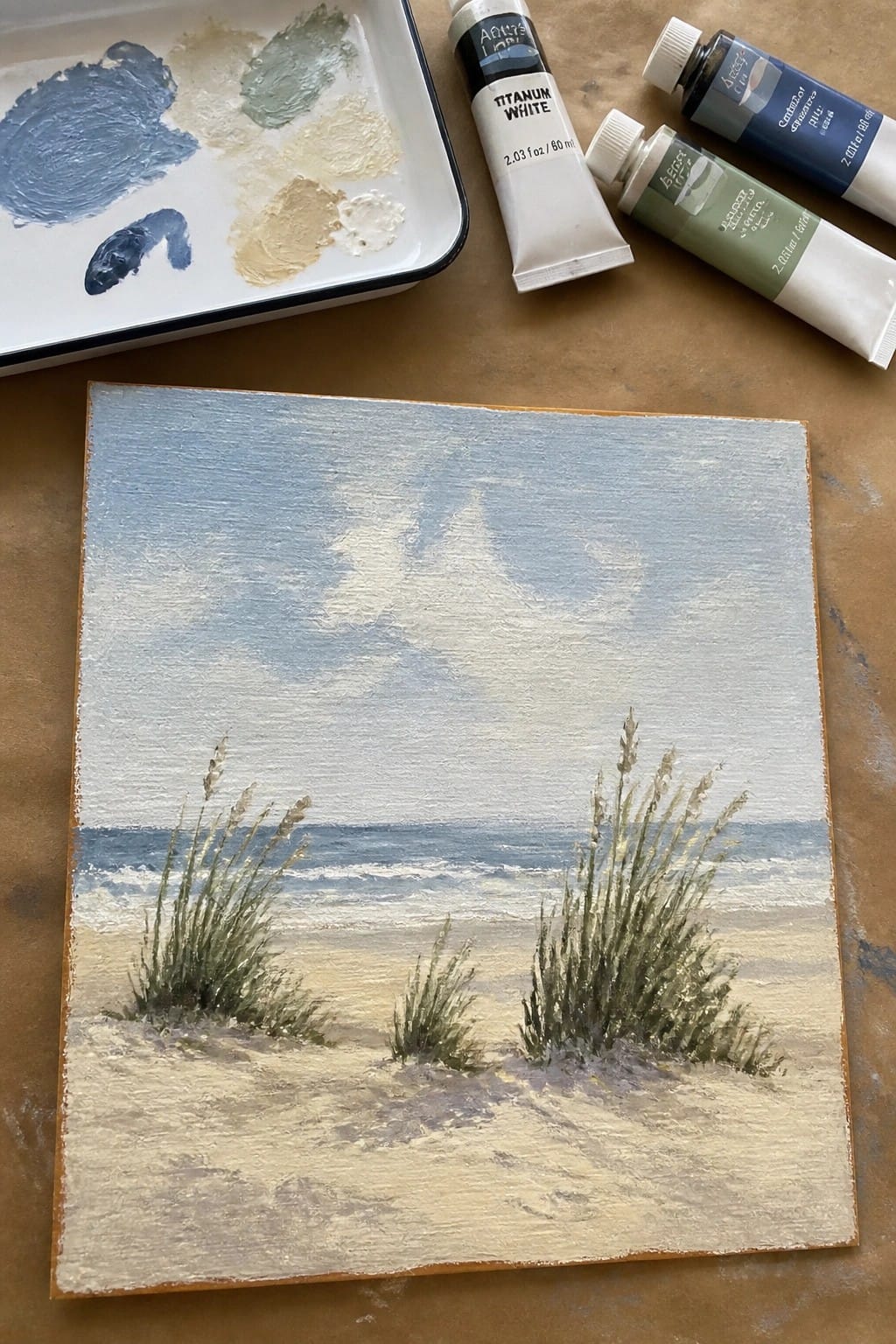 Beach painting with ocean waves and dune grass beside paint tubes and palette