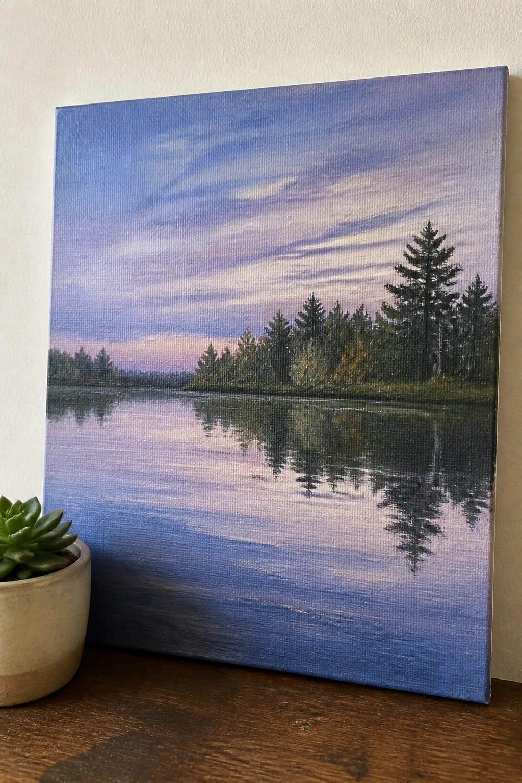 Landscape painting of reflective lake with pine trees under purple sky beside potted succulent.