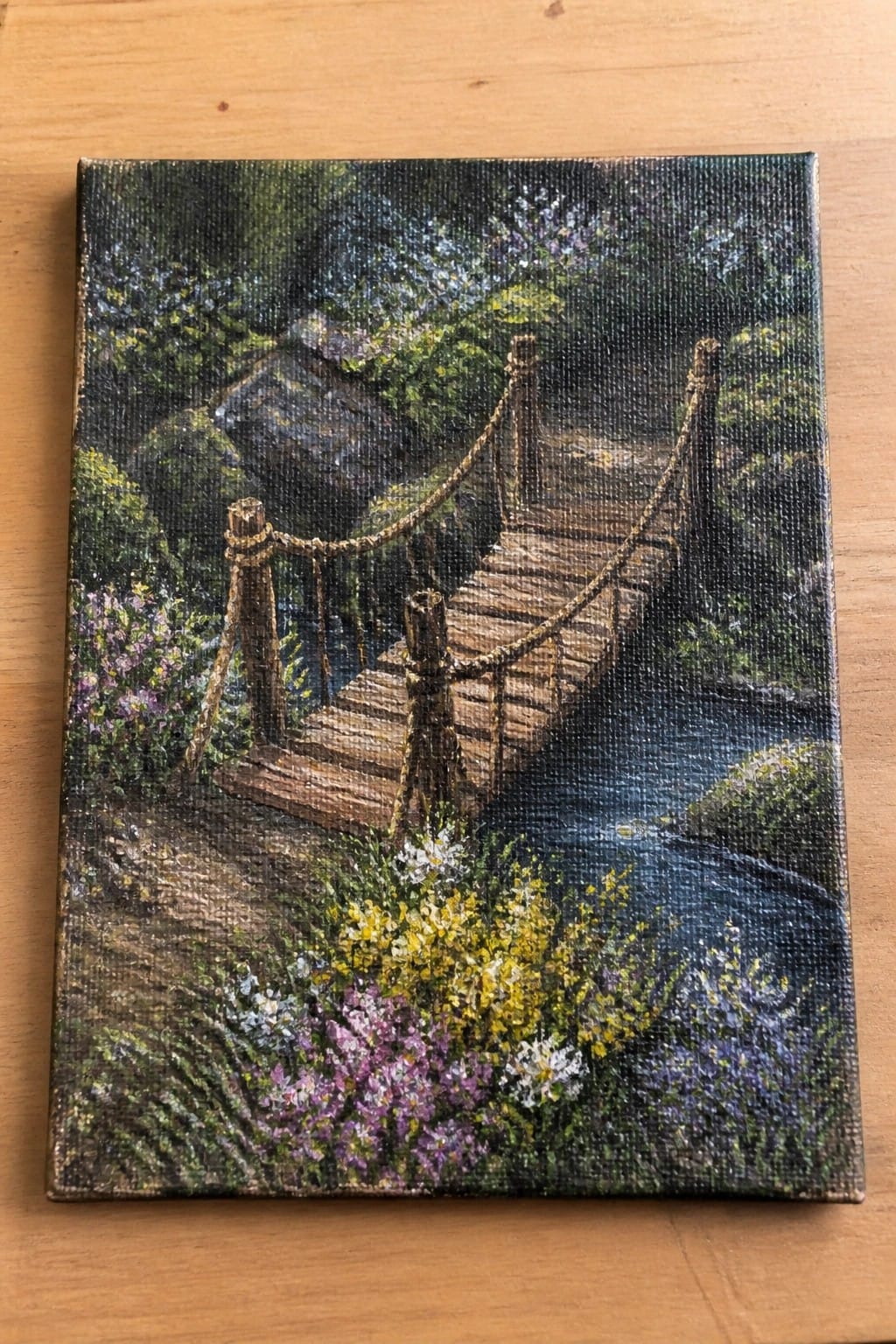 Wooden footbridge with rope railings spans a blue stream amid colorful wildflowers and trees