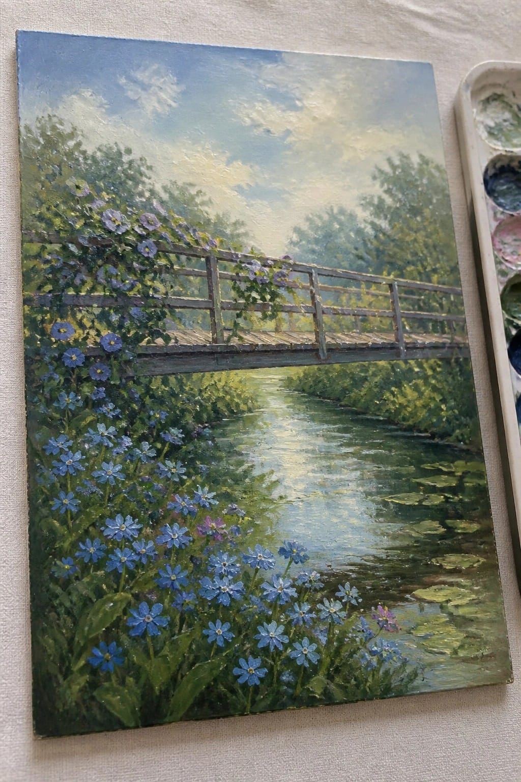 Oil painting of wooden bridge over stream amid blue flowers and lush greenery, palette nearby.