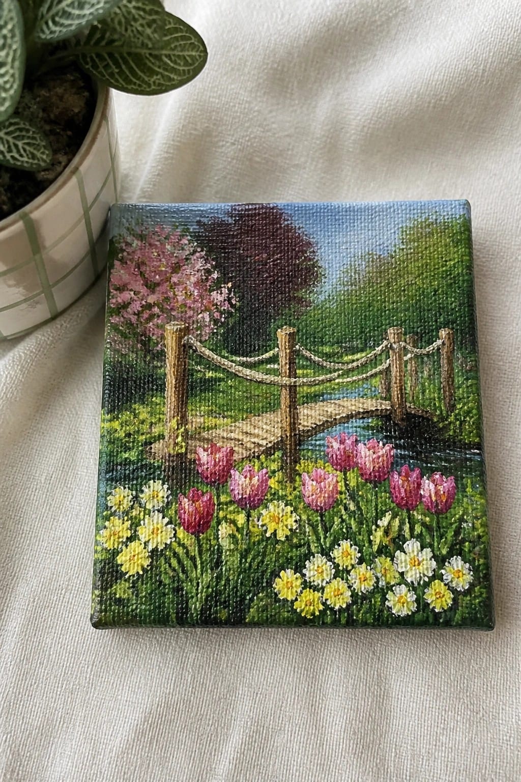 Small painting of tulips, bridge over stream, and blooming trees beside potted plant.