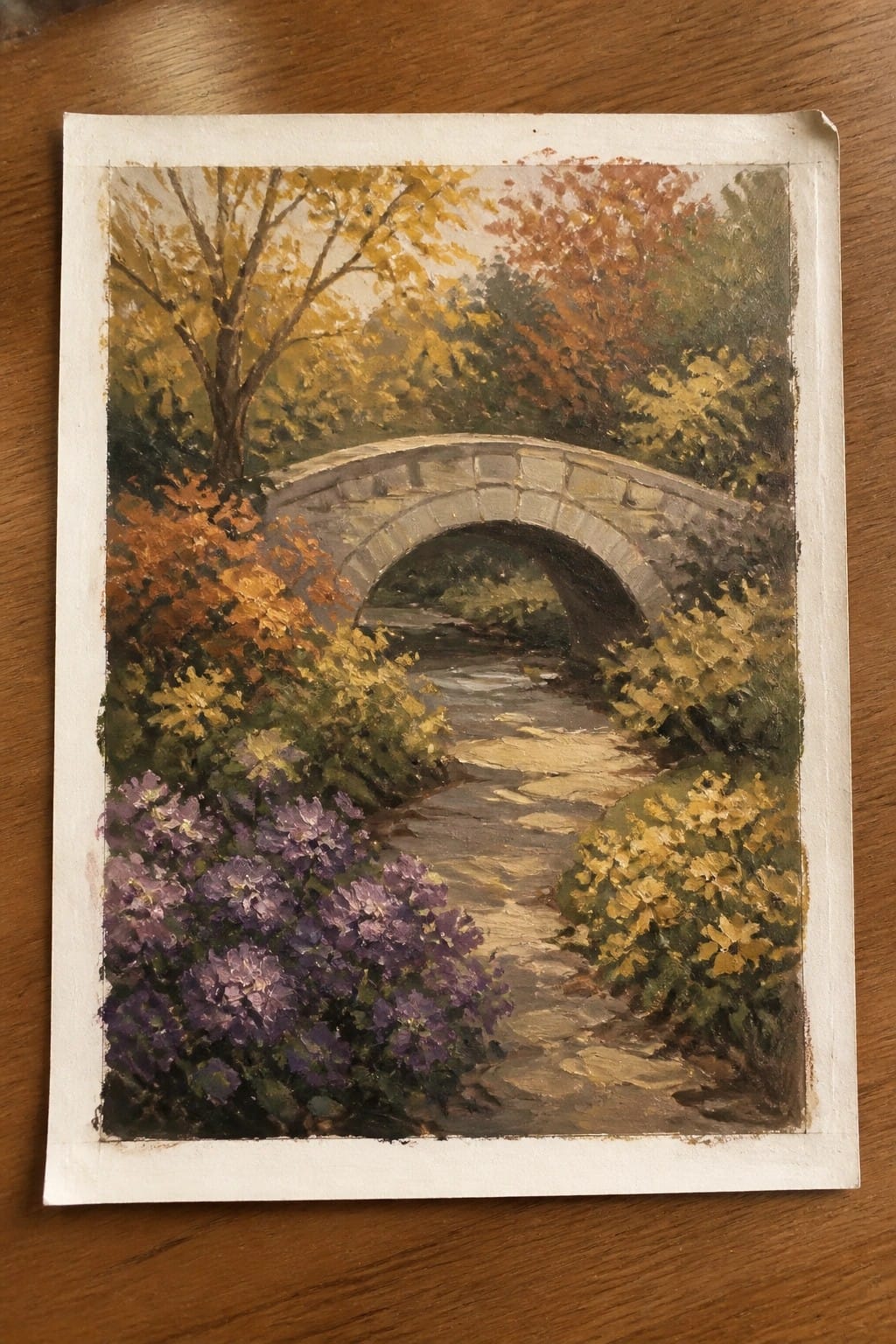 Stone bridge arches over stream amid autumn trees and purple flowers.
