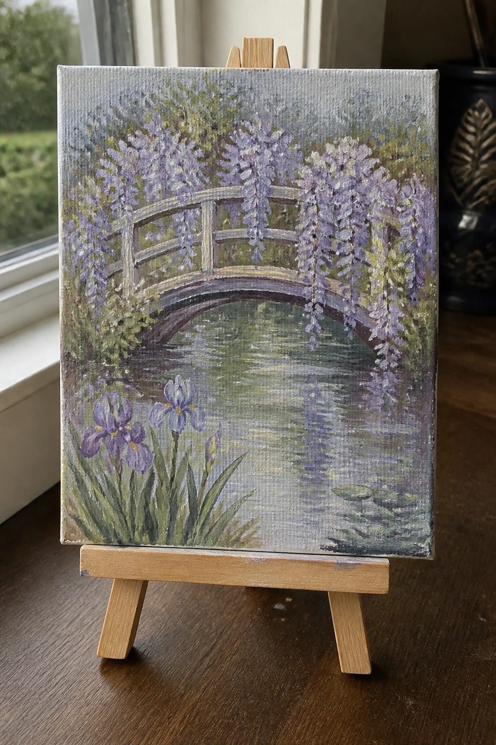 Small canvas on easel depicts arched bridge draped in purple wisteria over a pond with irises.