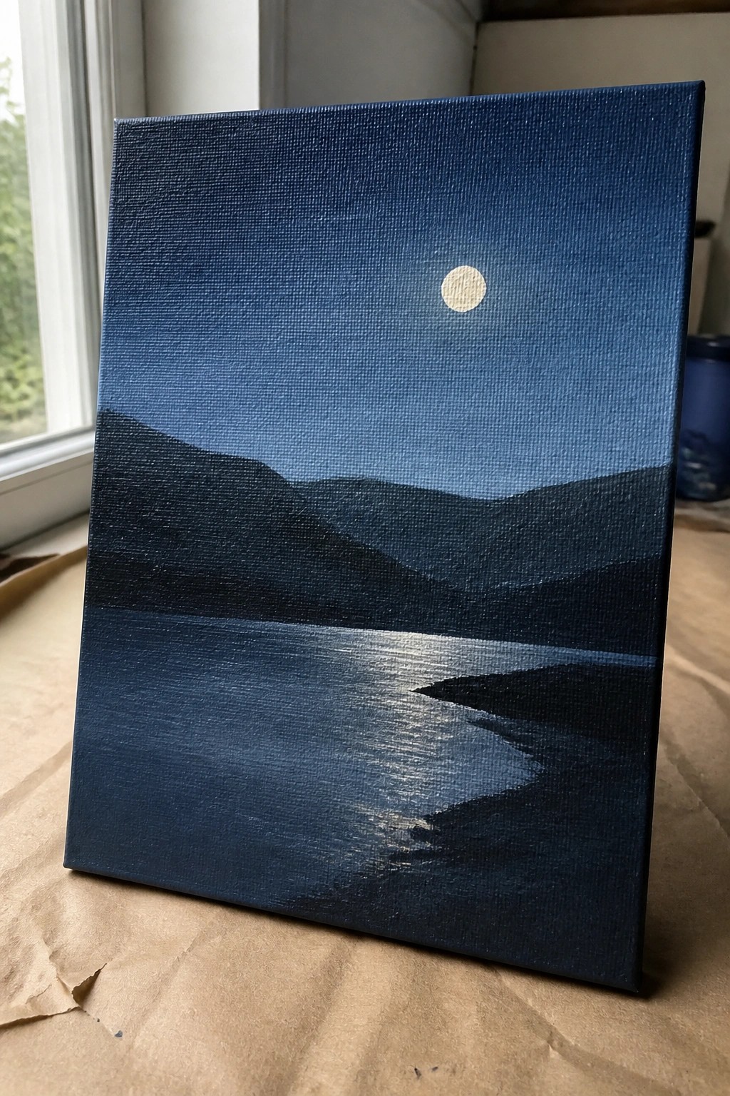 Dark blue painting of moon above layered hills with shimmering water reflection.