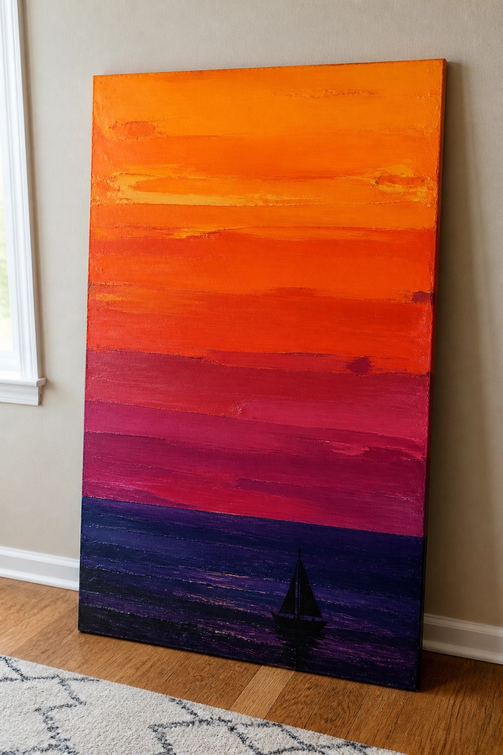 A rectangular oil painting of a sunset with horizontal stripes of orange, red, magenta, and purple, featuring a black sailboat silhouette on the lower section, leaning against a wall on a wooden floor.