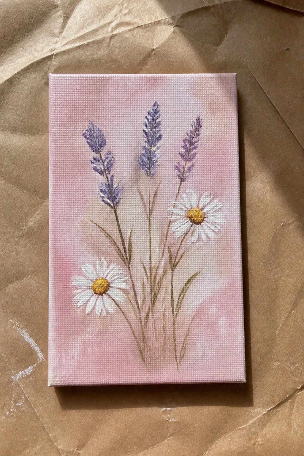 Small canvas with purple lavender and white daisies on pink background atop brown paper