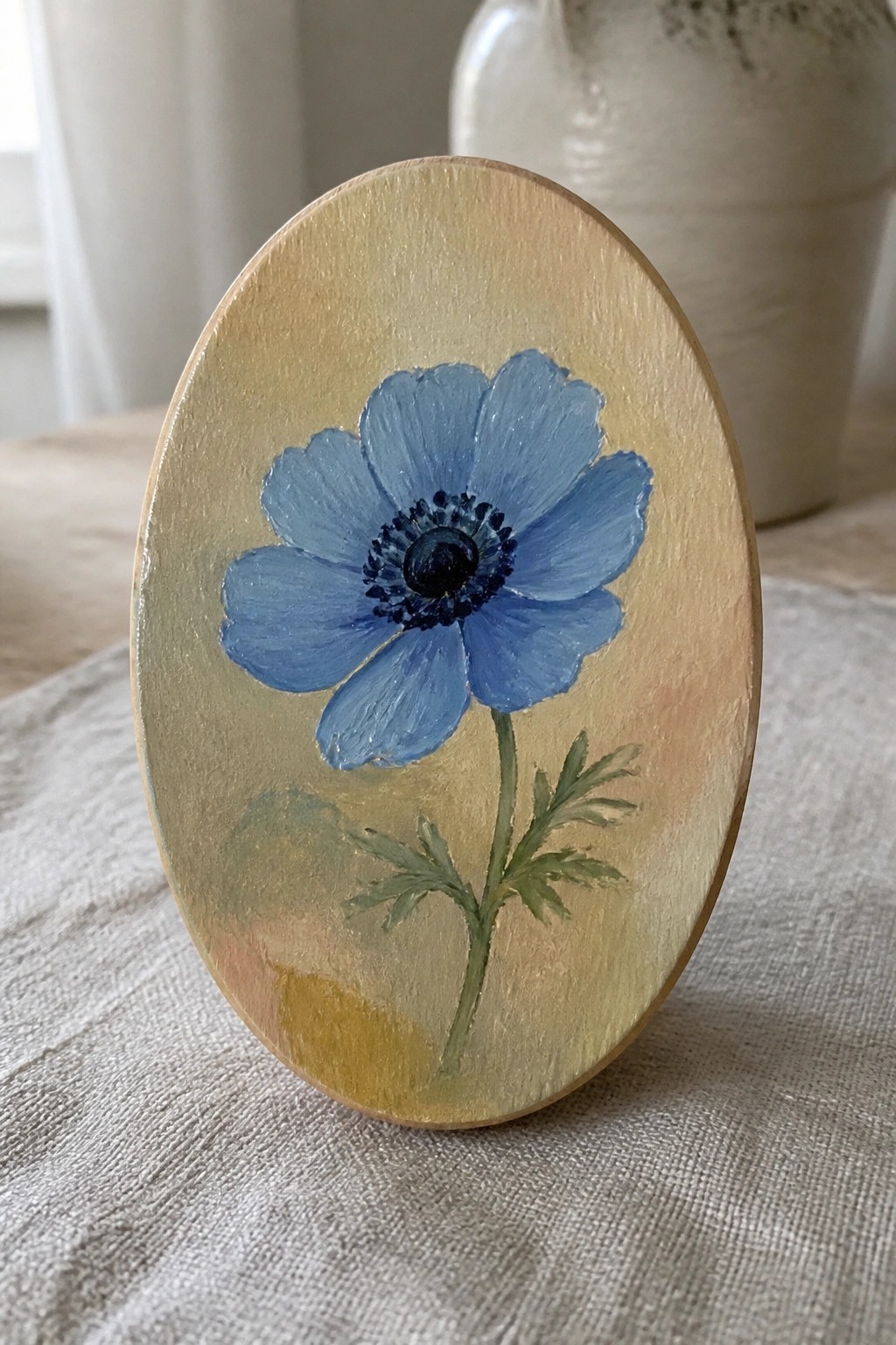 Oval wooden plaque with blue anemone flower and green stem on beige background.