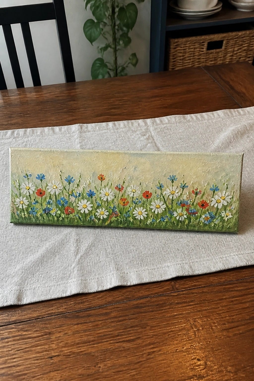 Horizontal canvas painting of colorful wildflowers on white cloth over wooden table