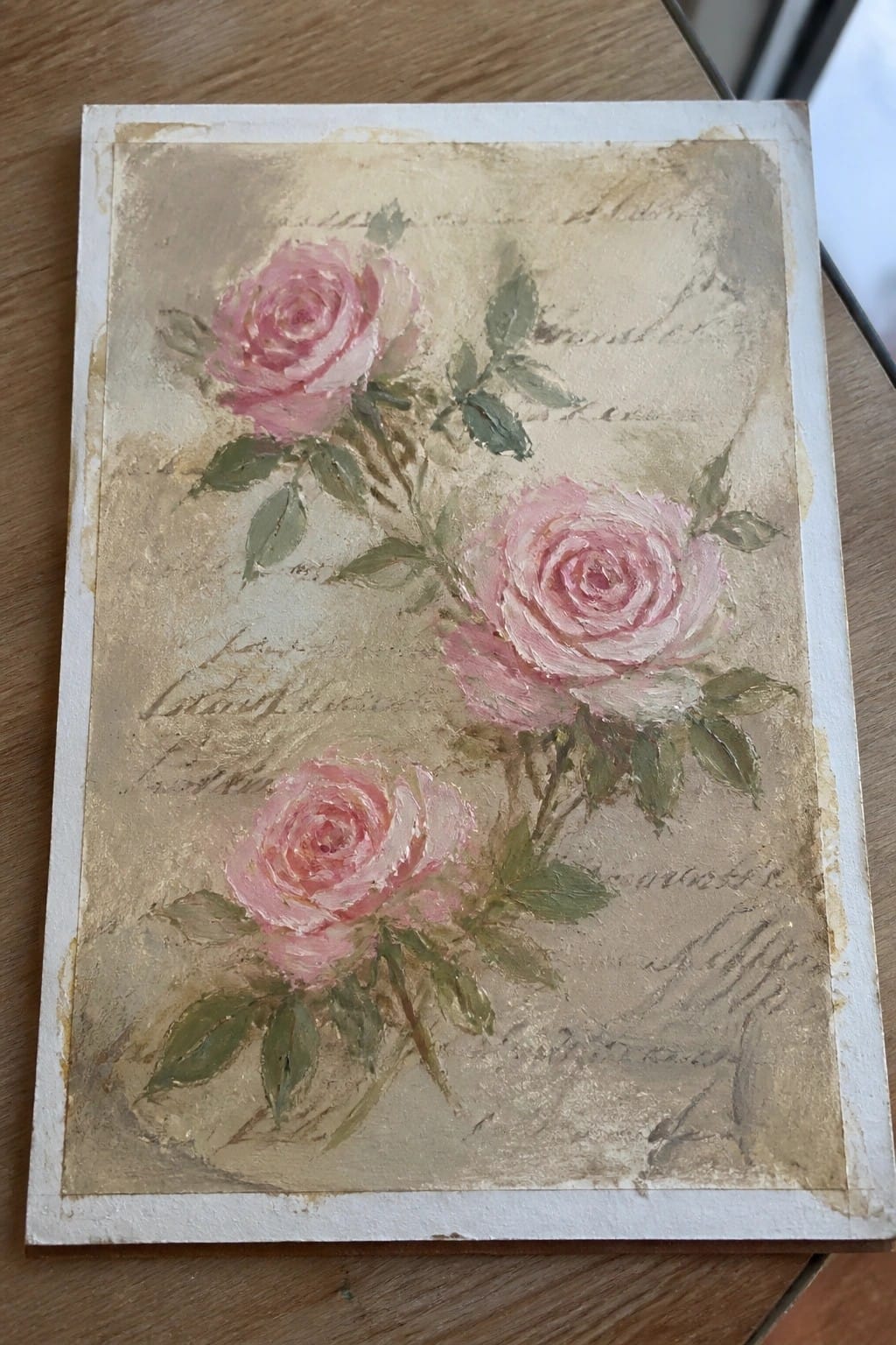 An oil painting of three pink roses with green leaves on a beige textured background with faint cursive writing.