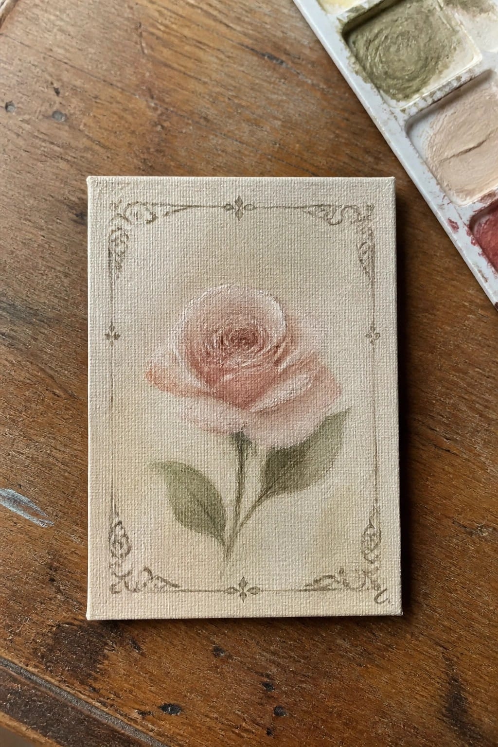 Small canvas with soft pink rose, ornate border, beside watercolor palette on wood