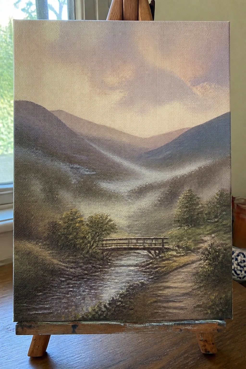 Realistic landscape painting on easel of misty mountains with bridge over stream