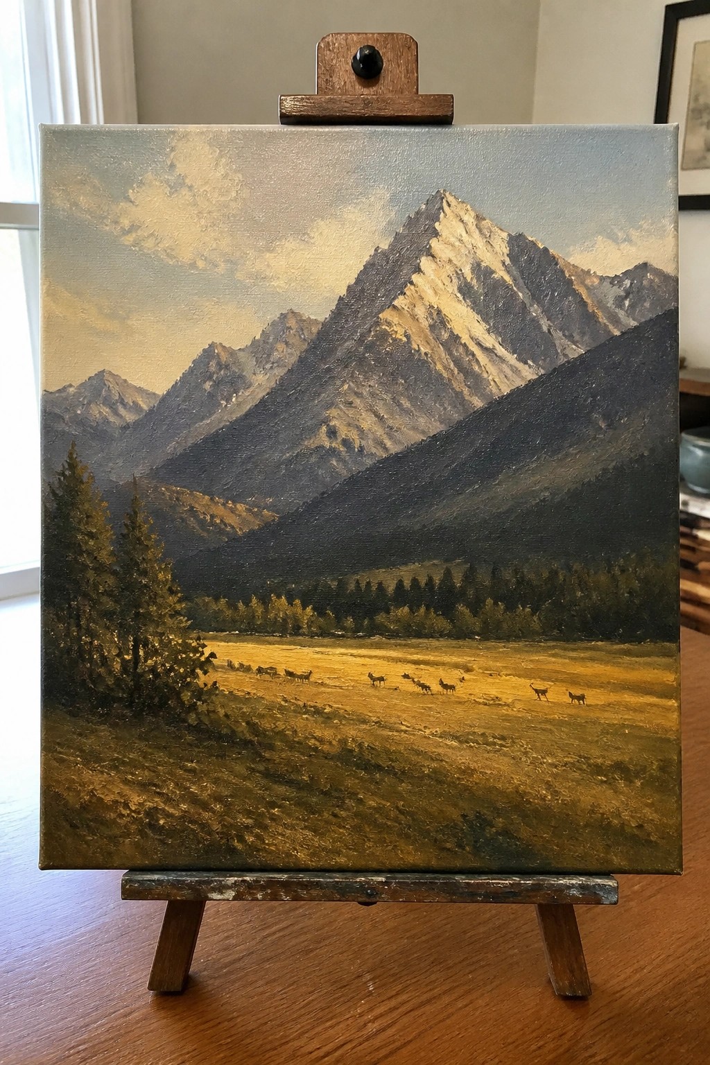Realistic oil painting on easel of snow-capped mountains above golden meadow with deer