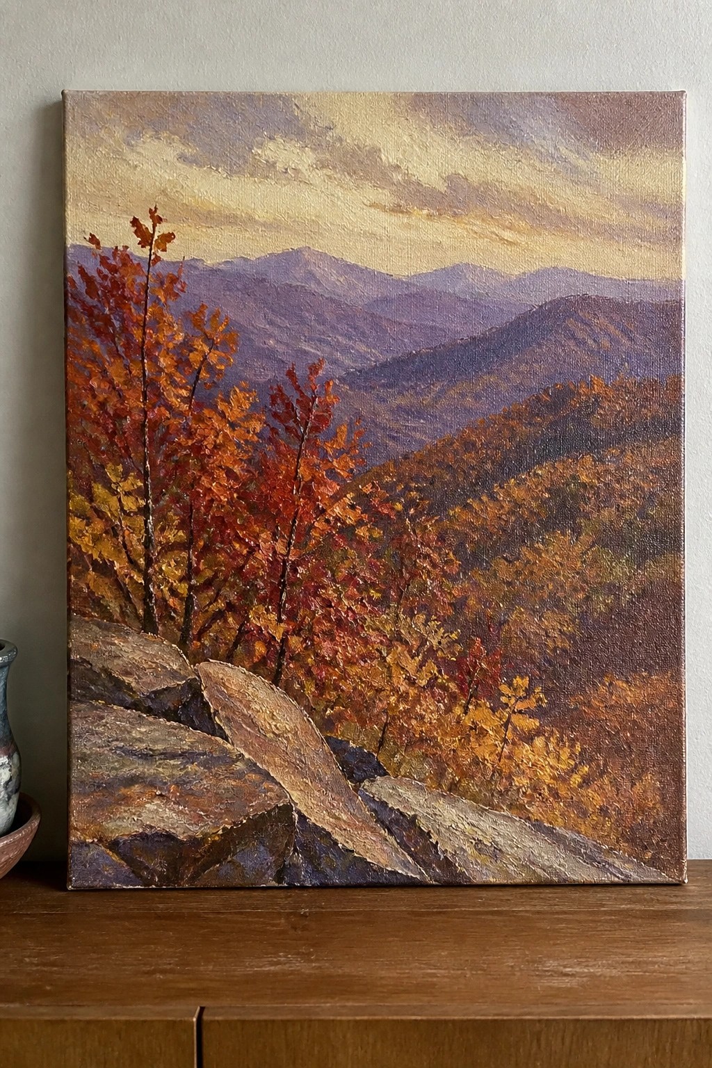 An oil painting of a mountain landscape with rocky foreground, red and orange autumn trees on the left, and layered purple mountains under a golden sky.