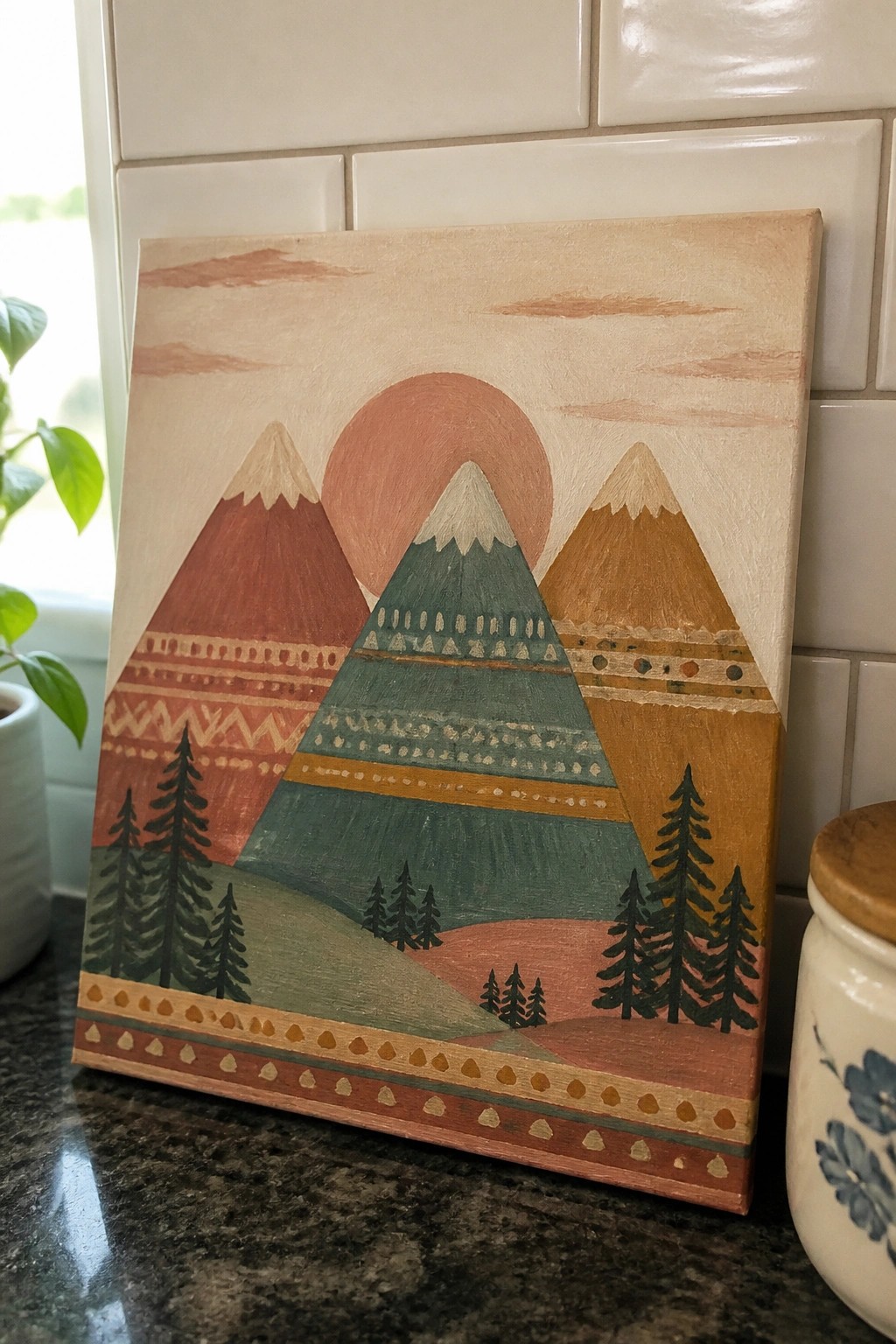 Folk-style canvas painting of patterned mountains, red sun, and pine trees in earthy tones.