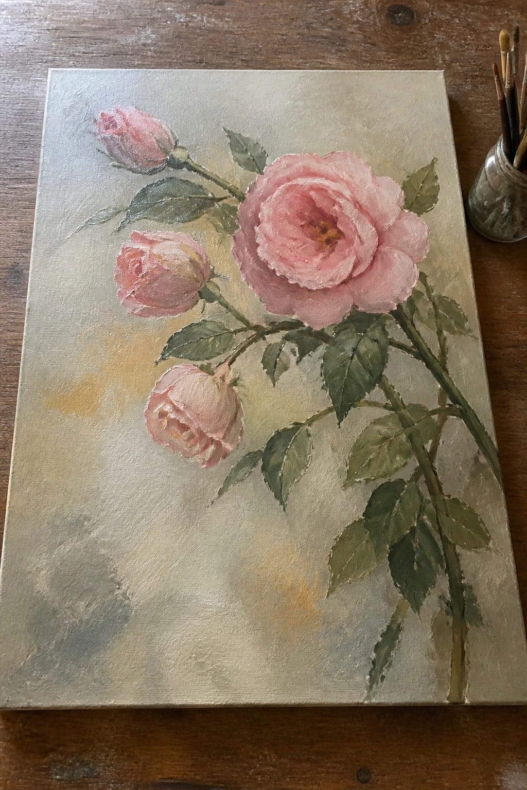Canvas with pink roses painting beside paintbrushes in glass jar on wood.