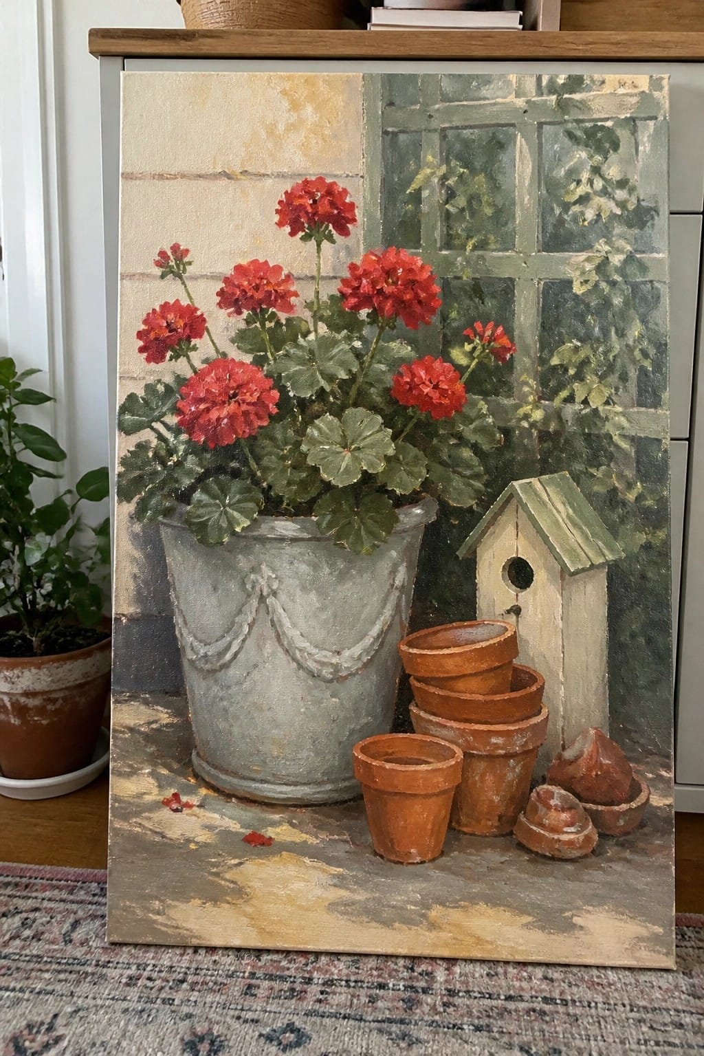 Still life painting of red geraniums in metal pot beside terracotta pots and birdhouse.