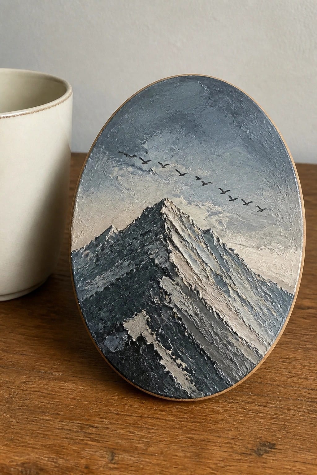 Thickly textured round painting of mountain with birds beside white cup on wood table