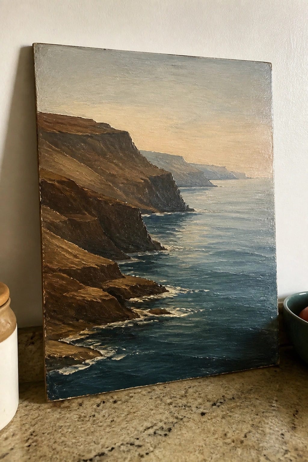 Oil painting of brown cliffs by blue ocean waves on marble countertop with jars.