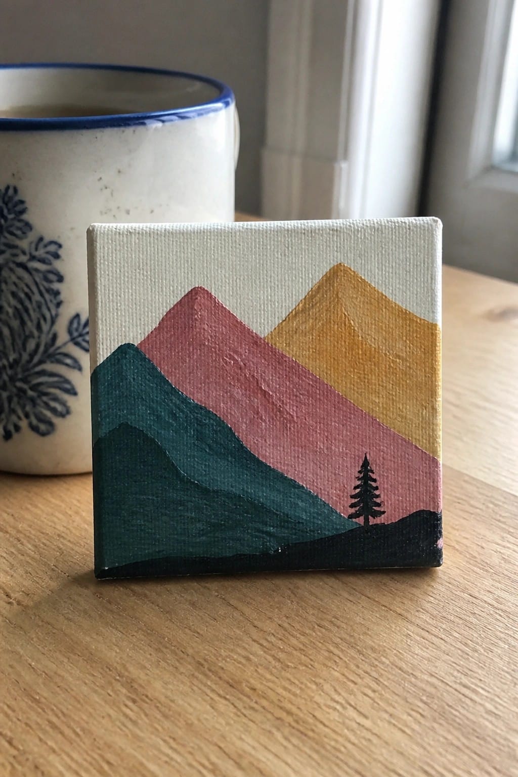 Mini canvas with layered teal, pink, and gold mountains and black pine tree on wood table