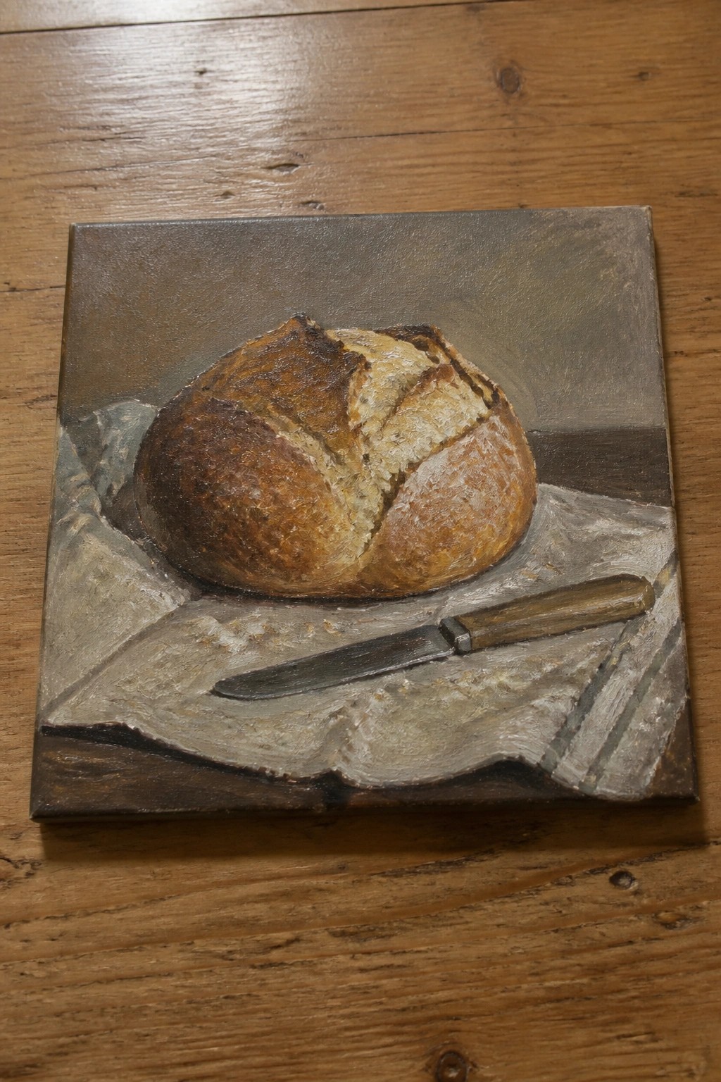 Crusty round bread loaf on cloth beside knife in still life painting.