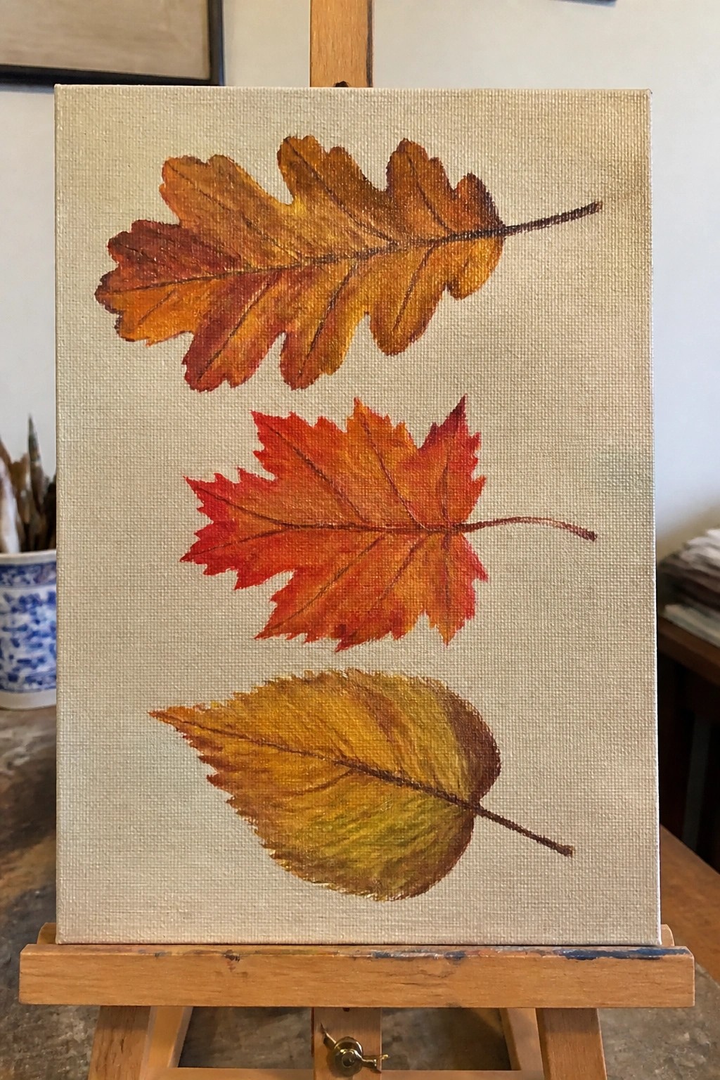 Three autumn leaves painted in orange, red, and yellow on canvas easel.