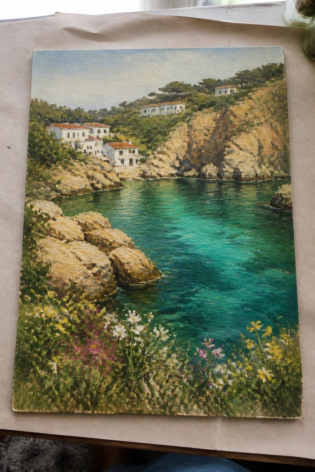 Coastal painting of turquoise cove, rocky cliffs, white houses, and wildflowers.
