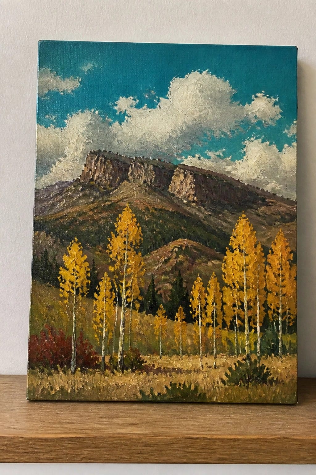 Oil painting of a rocky mountain peak with yellow aspen trees in front and a blue sky with white clouds.