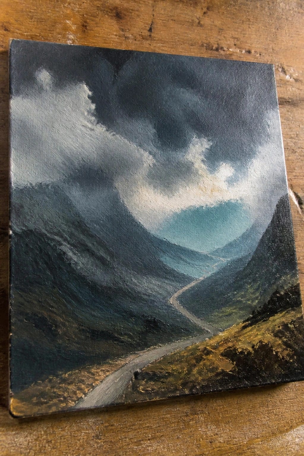Dramatic oil painting of winding road through dark mountains under stormy cloudy sky with light.