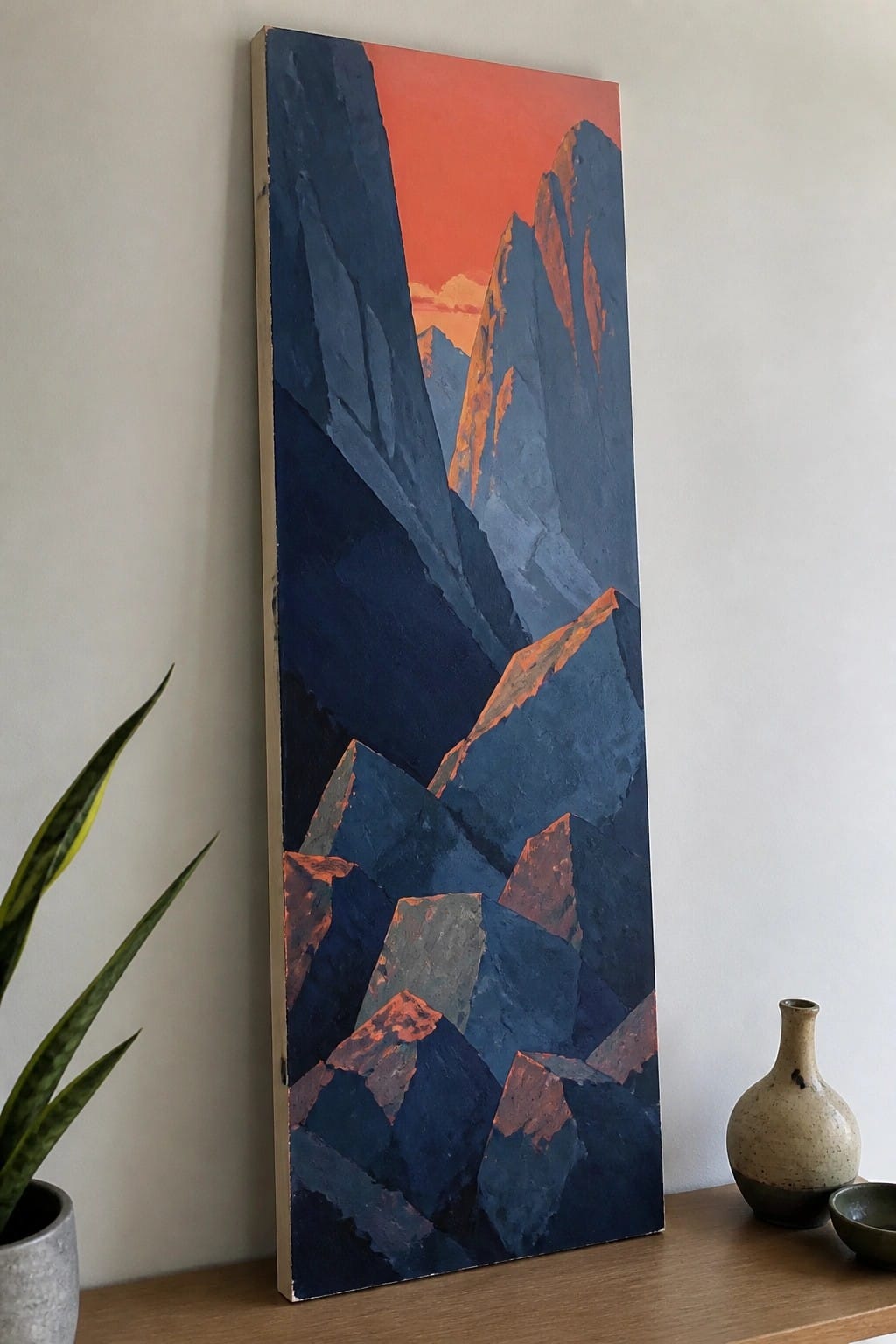 A tall vertical oil painting of angular dark blue mountains with orange highlights against a red sky, placed on a wooden surface next to a plant and pottery.