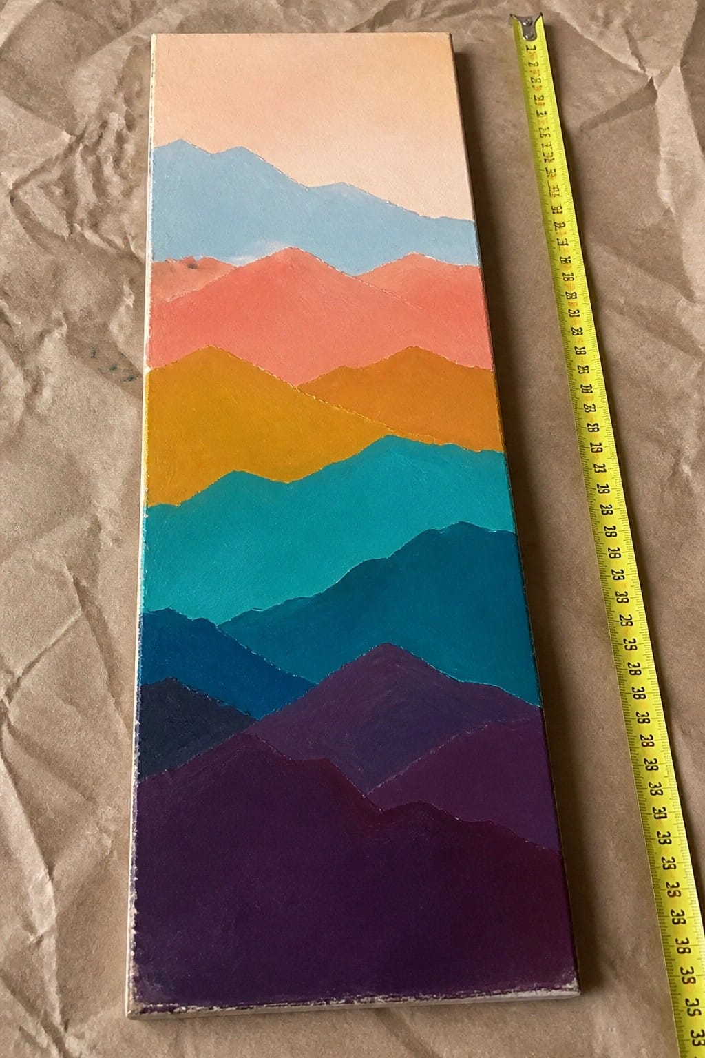 Vertical colorful mountain landscape painting beside yellow measuring tape on brown paper.