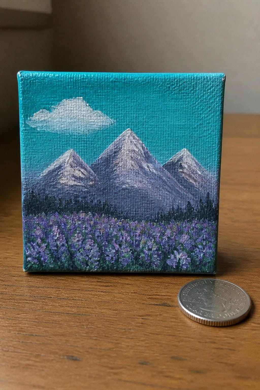 Small square painting of snow-capped mountains above purple flowers beside a quarter coin