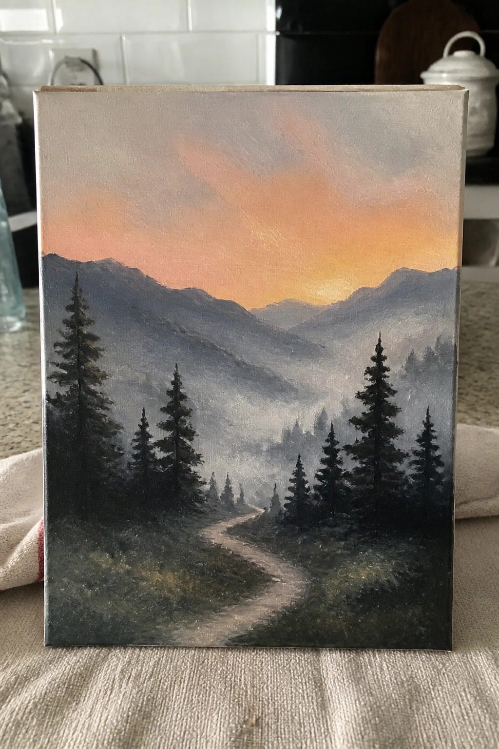 Canvas painting of misty mountains, winding path through pine trees under orange sky.