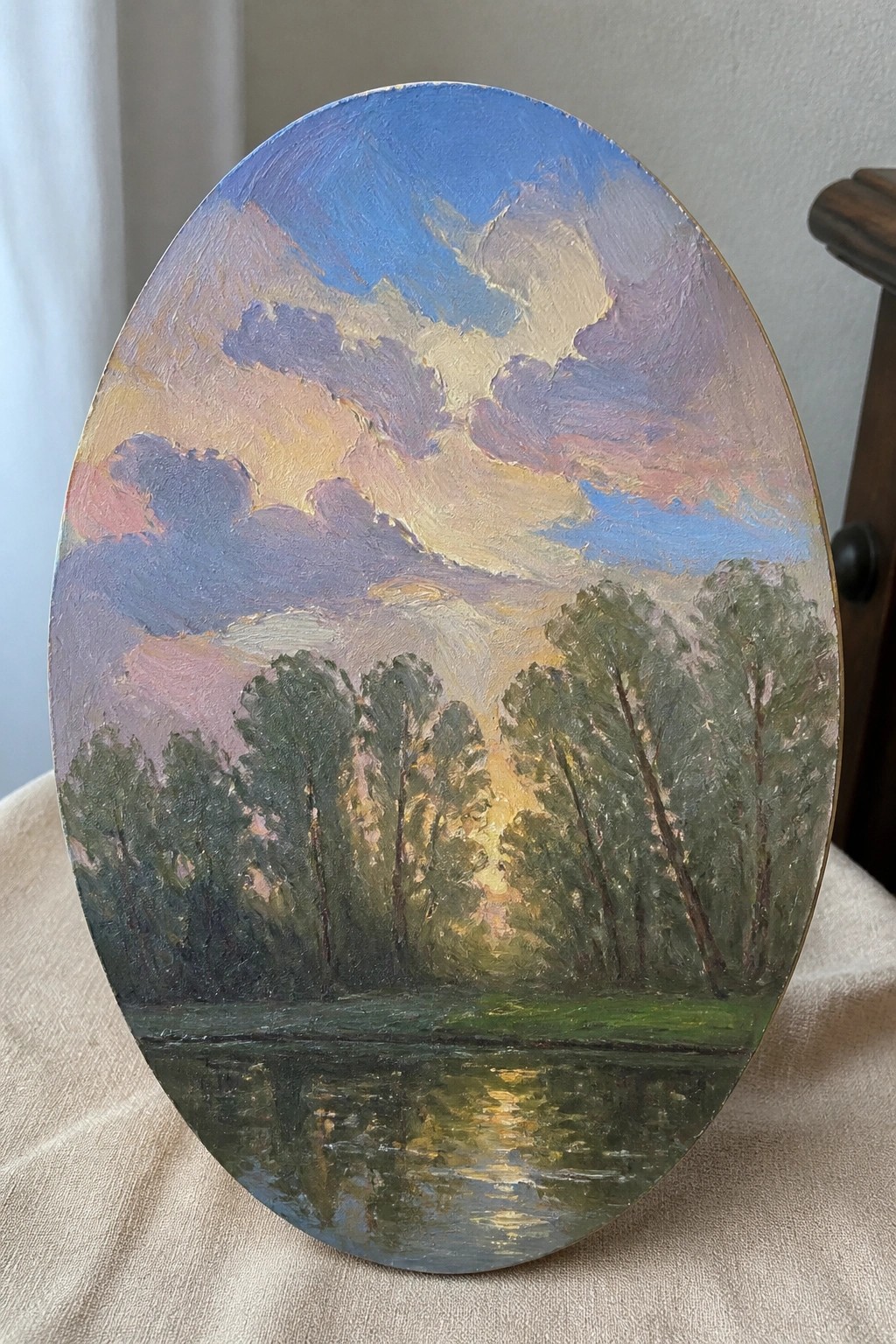 Oval oil painting of sunset clouds over trees reflected in calm water.