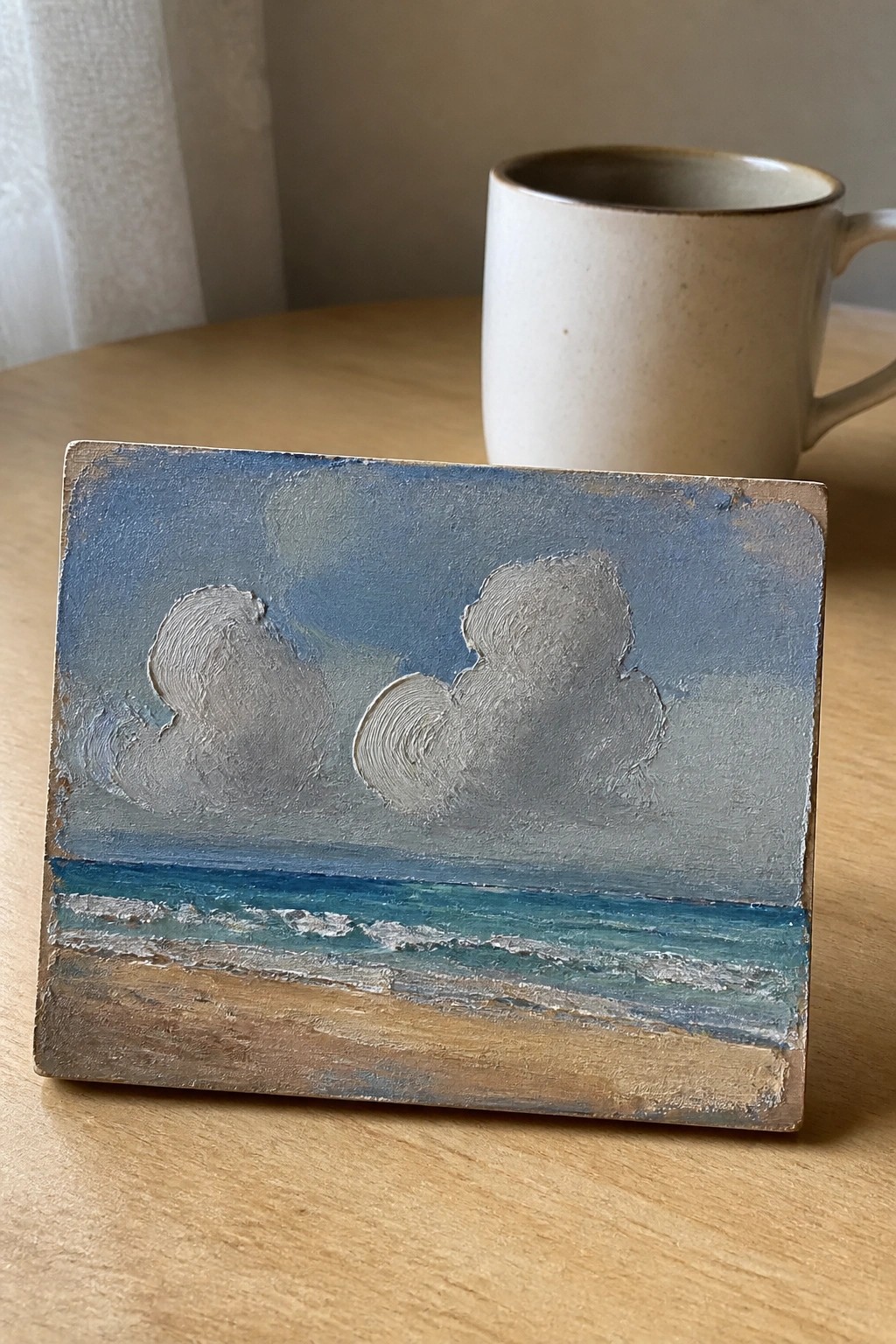 A small rectangular oil painting on wood shows a blue sky with textured white clouds above a strip of turquoise water with wave lines and a sandy beach below.