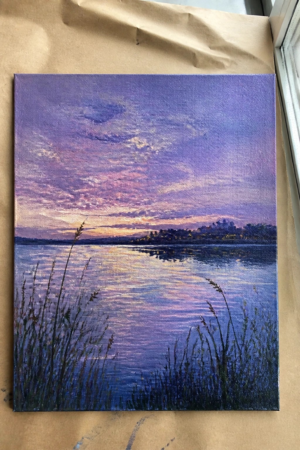 Purple-pink sunset painting over reflective lake with silhouetted foreground grass on canvas