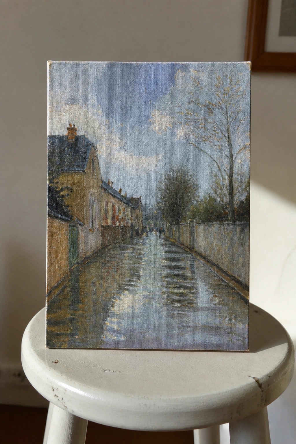Oil painting of wet village street with reflections propped on white stool.